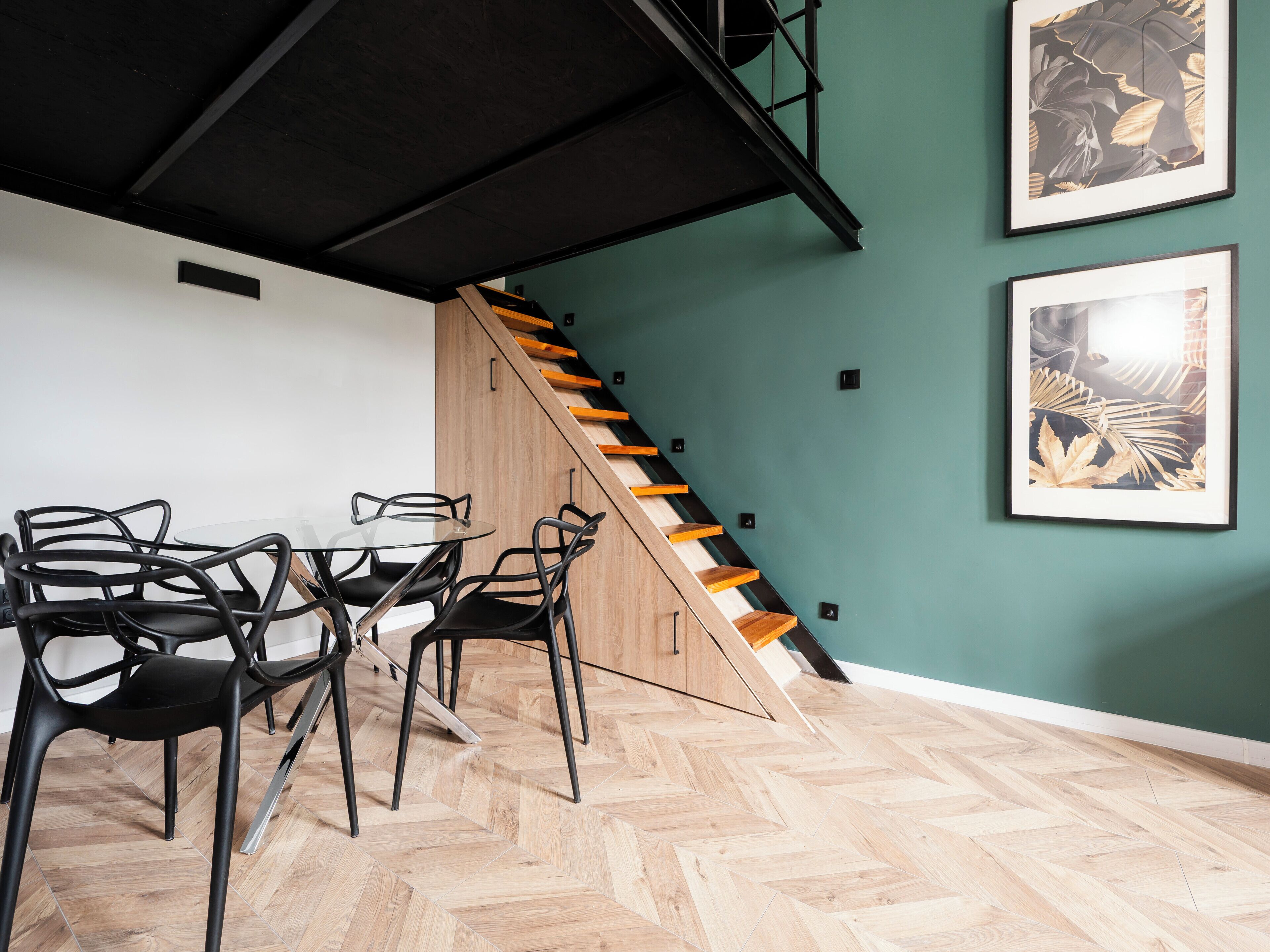 A dining area with a table and chairs, a staircase, and two framed artworks on the wall.