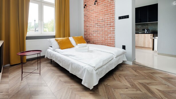 A modern bedroom with a brick accent wall, a bed with white bedding, a small table, and a view of a kitchen. - Nawrot City Center Studio (Łódź)
