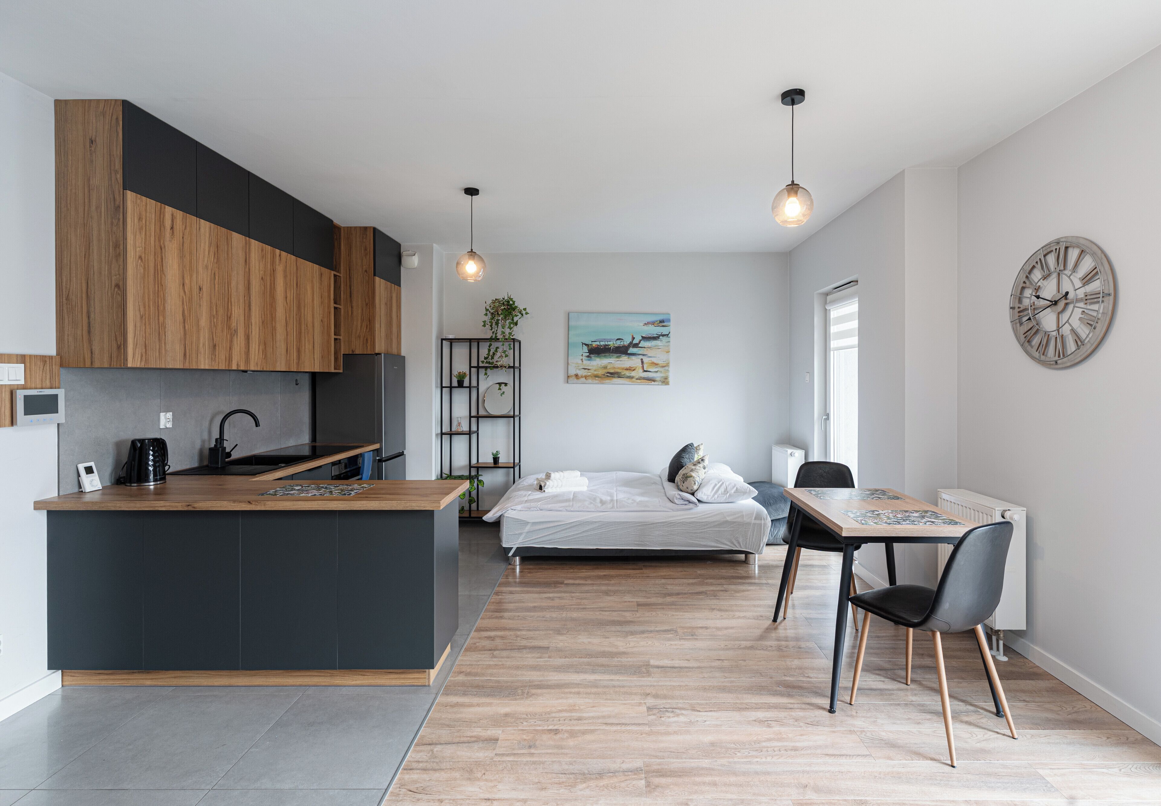 Une cuisine moderne avec un îlot central, un coin repas avec une table et des chaises, un lit avec vue et une horloge au mur.