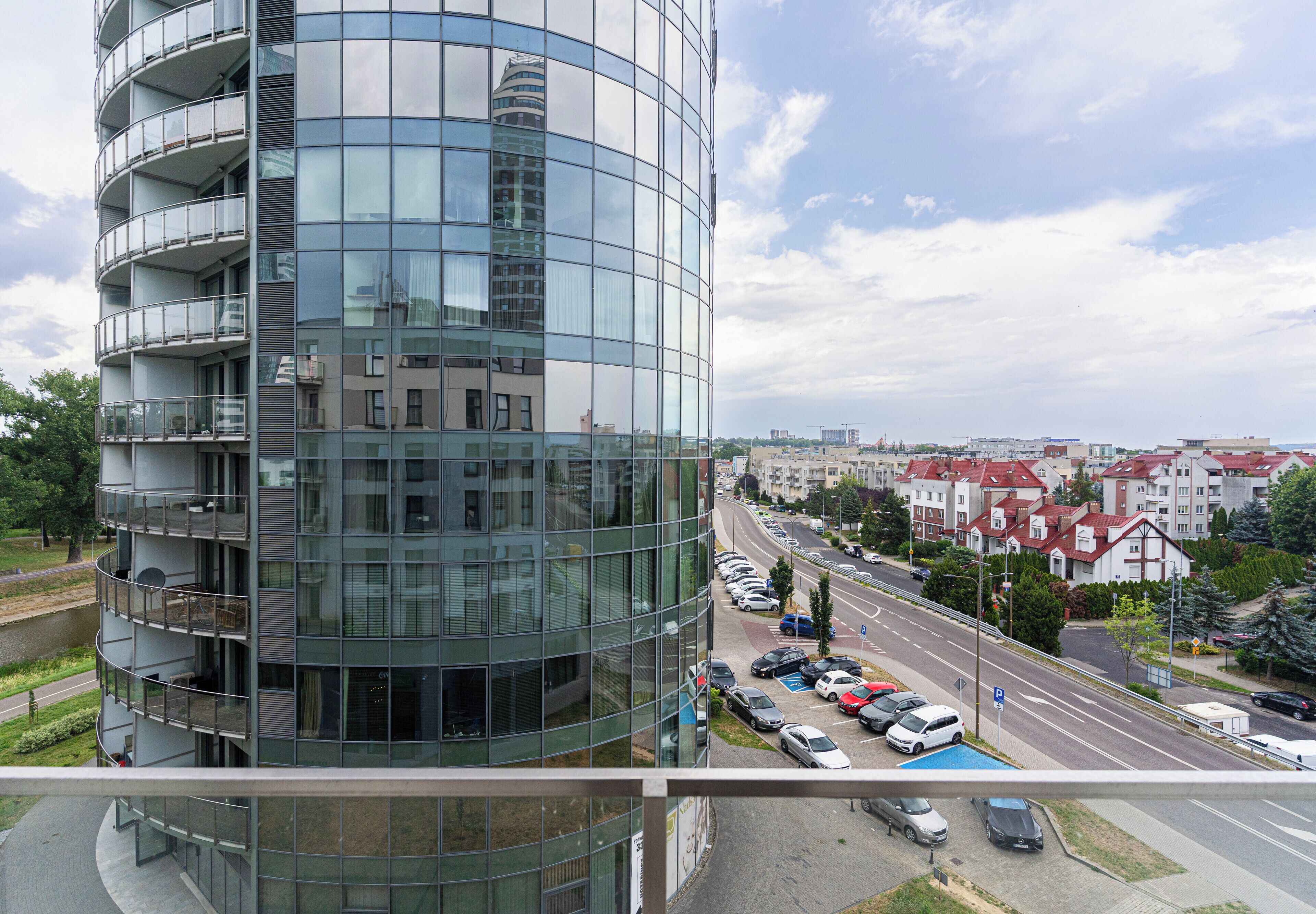 A modern multi-story building with a curved glass facade, a parking lot with cars, and a view of a residential area with red-roofed houses.