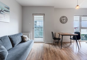 A modern living room with a grey sofa, a dining table with two chairs, a clock on the wall, and a view through a glass door. - Podwisłocze Modern Apartment (Rzeszów)