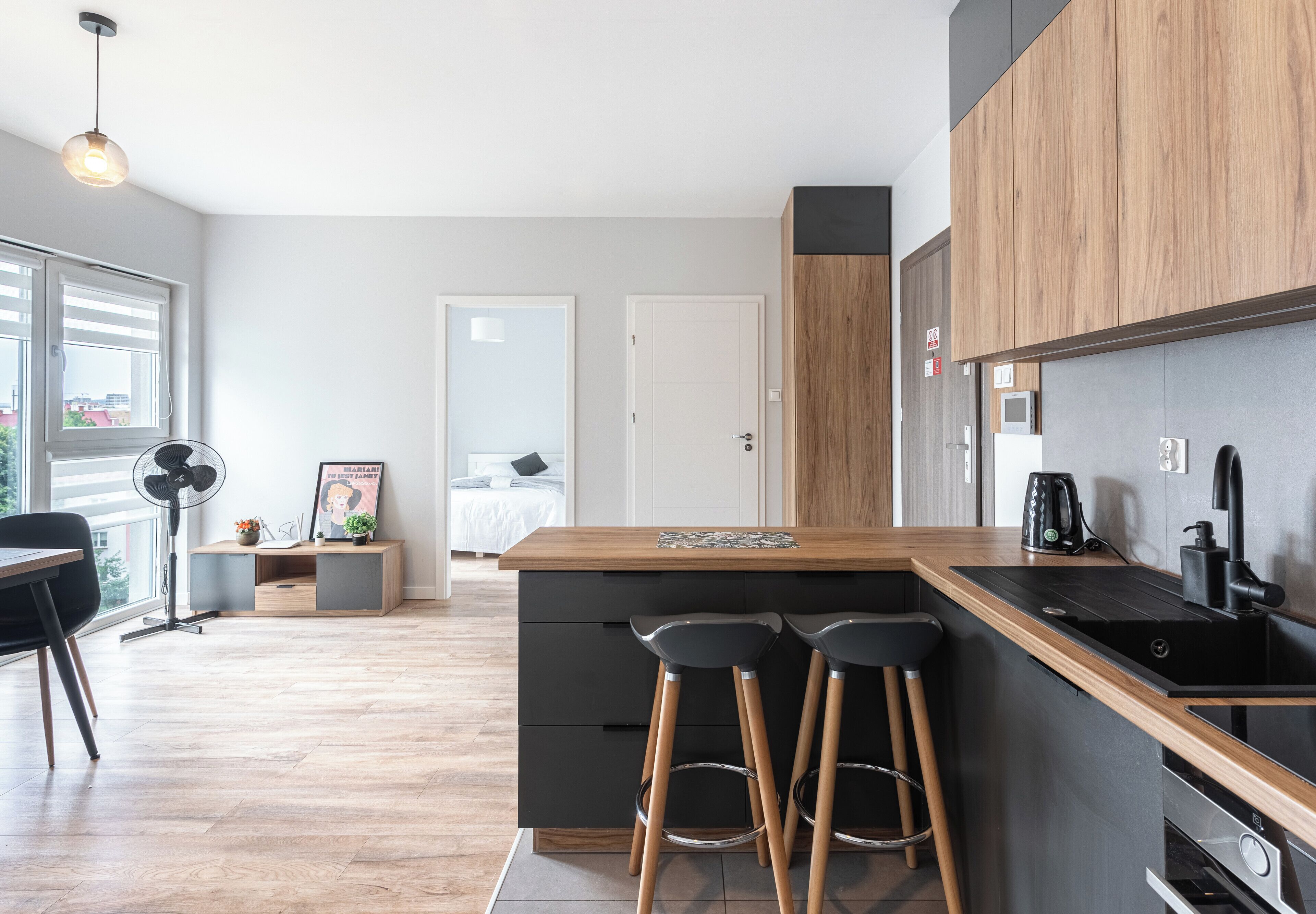 Une cuisine moderne avec un plan de travail en bois, des tabourets de bar noirs et, au fond, une vue sur la chambre à coucher.