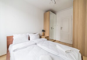 Uma cama bem arrumada com lençóis brancos, cabeceira de madeira, mesa de cabeceira com abajur e um espelho grande.