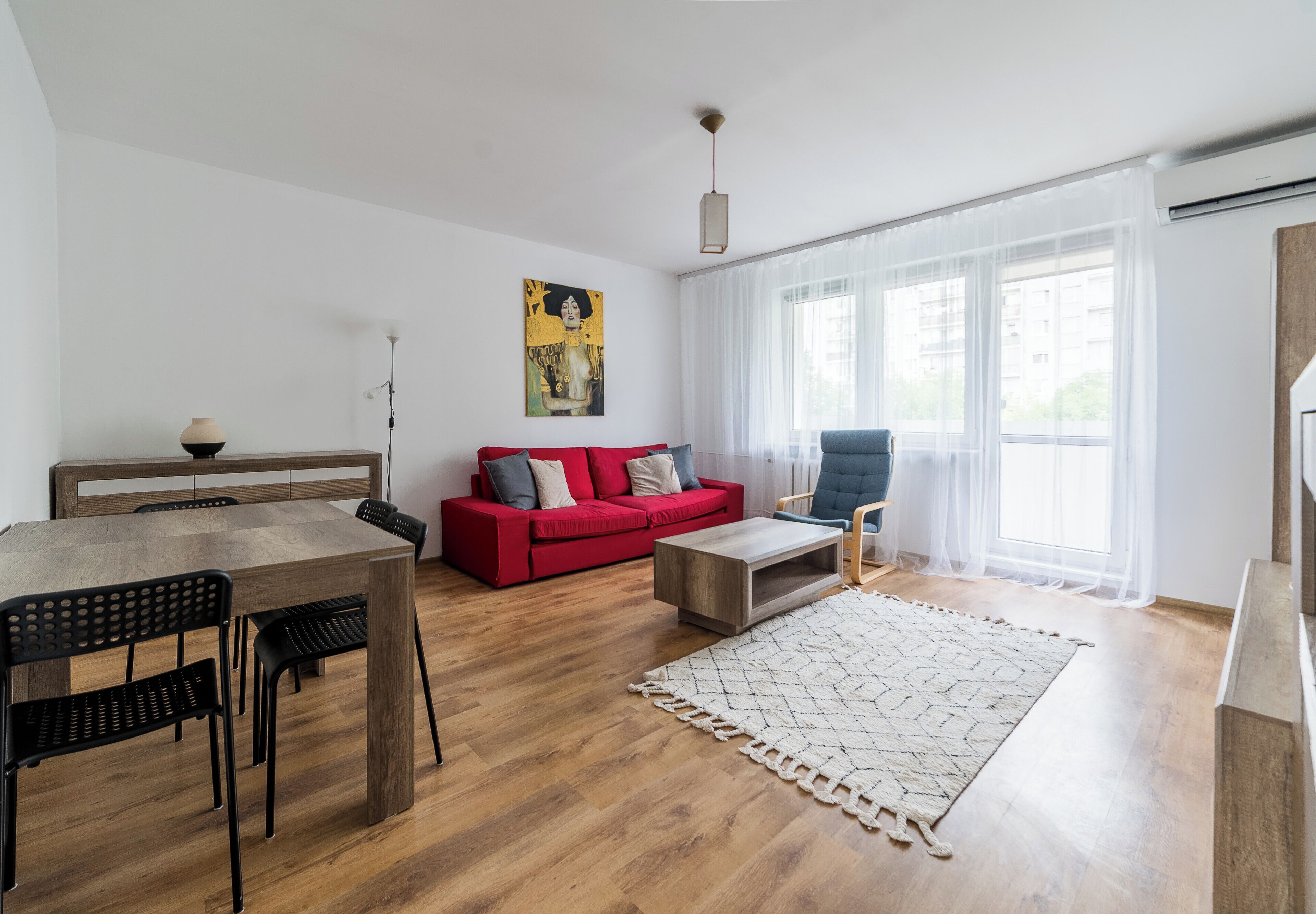 A modern living room with a dining area, a red sofa, a blue armchair, and a large window with sheer curtains.