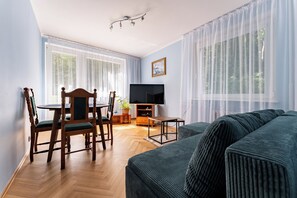 A living room with a sofa, a dining table with chairs, a TV, and large windows with sheer curtains. - Spadzista Comfort Apartment (Gdańsk)