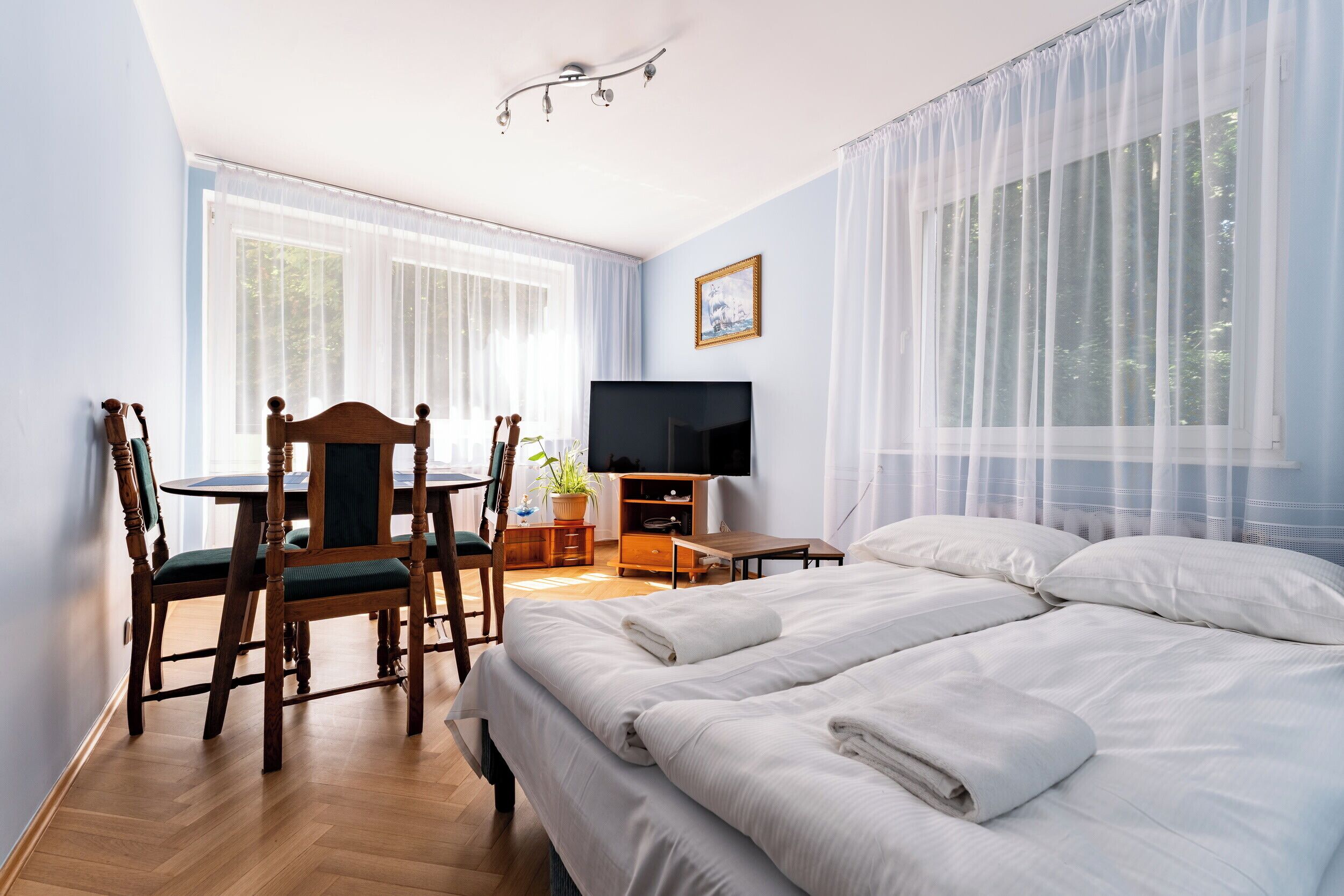 A hotel room with a bed, a desk with a chair, a television, and a window with sheer curtains.