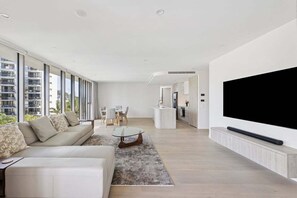 A modern living room with a sofa, coffee table, and TV. There is a kitchen area with a sink and cabinets. - 3BRM Oceanview Elegance (Gold Coast)