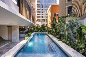 Ein Pool mit modernem Design, umgeben von üppiger tropischer Vegetation und an ein mehrgeschossiges Gebäude mit Balkonen angrenzend.