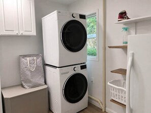 A laundry room with two stacked washing machines, a drying rack, a basket, and a shelf with a shoe and a spray bottle.