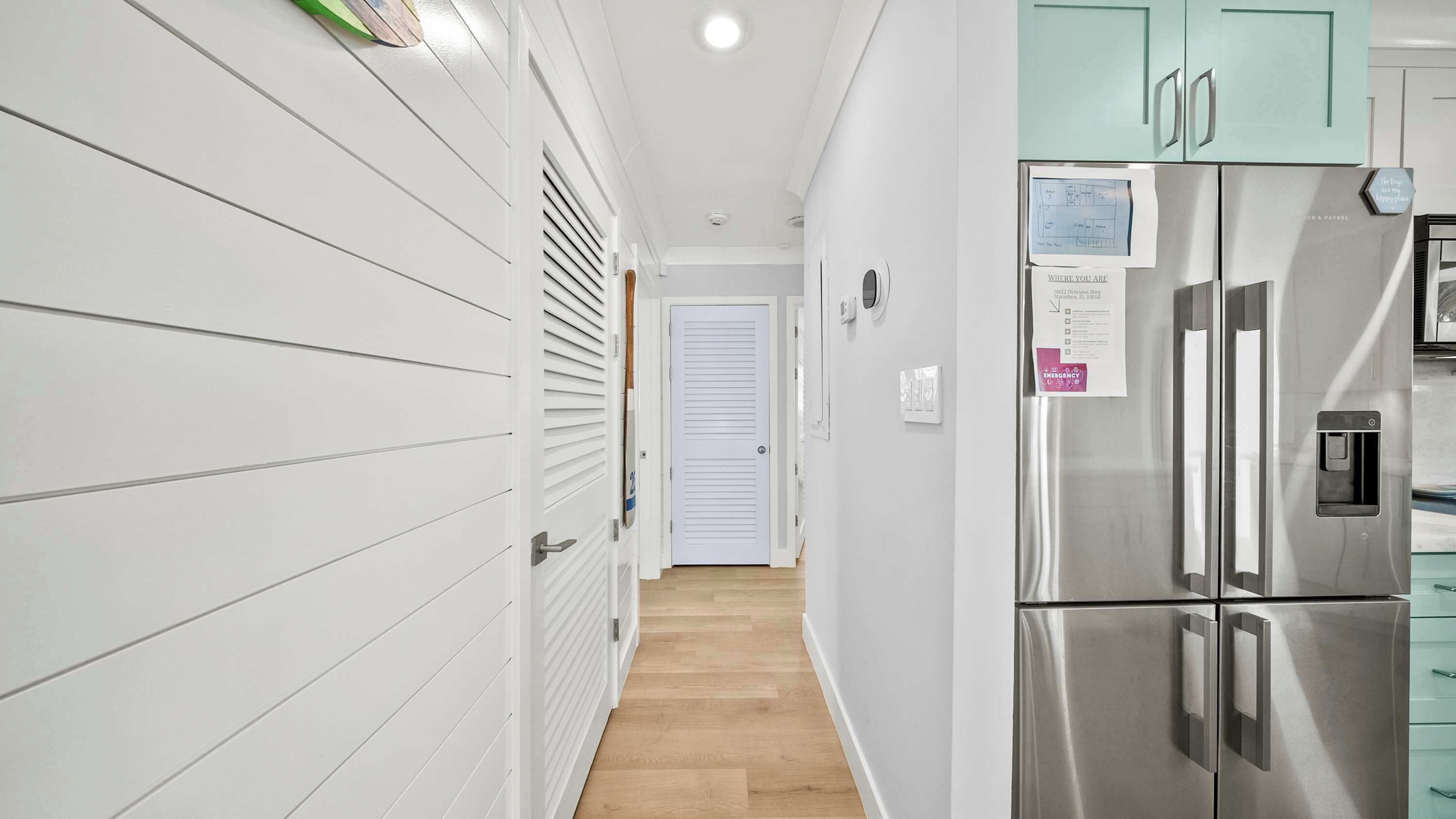 A hallway with white walls, a wooden floor, a stainless steel refrigerator, and a door with a number.