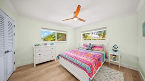 Ein Schlafzimmer mit einem Bett, einer Kommode, einem Nachttisch, einem Deckenventilator und Fenstern mit Blick auf Grünflächen.