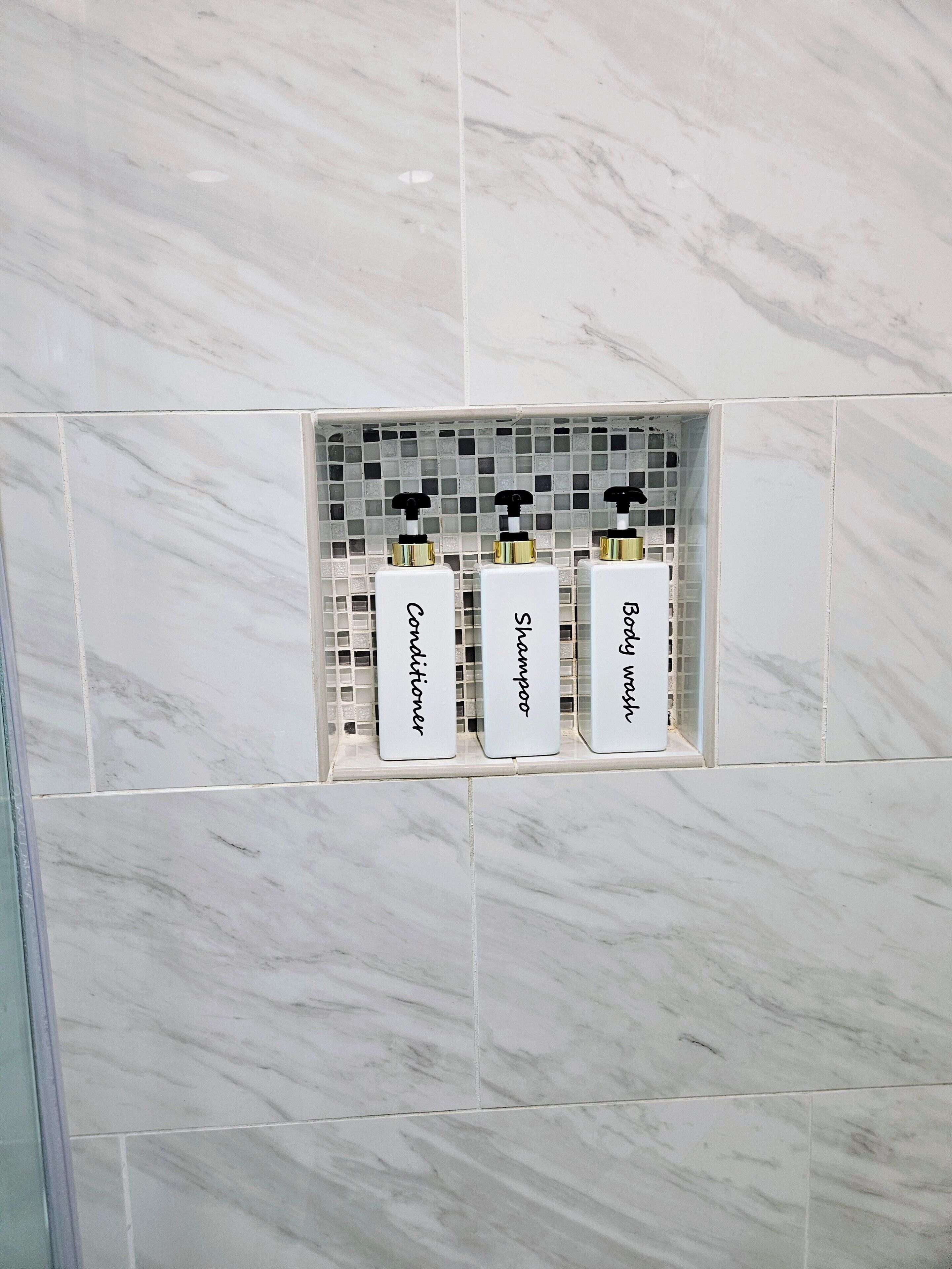 Ein Badezimmer mit gefliestem Boden und einer Ablage mit drei Flaschen: Conditioner, Shampoo und Duschgel.