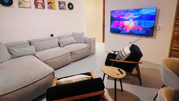 Un salon comprenant un canapé, une petite table, un fauteuil, un ventilateur de plafond et un téléviseur affichant une image de paysage.