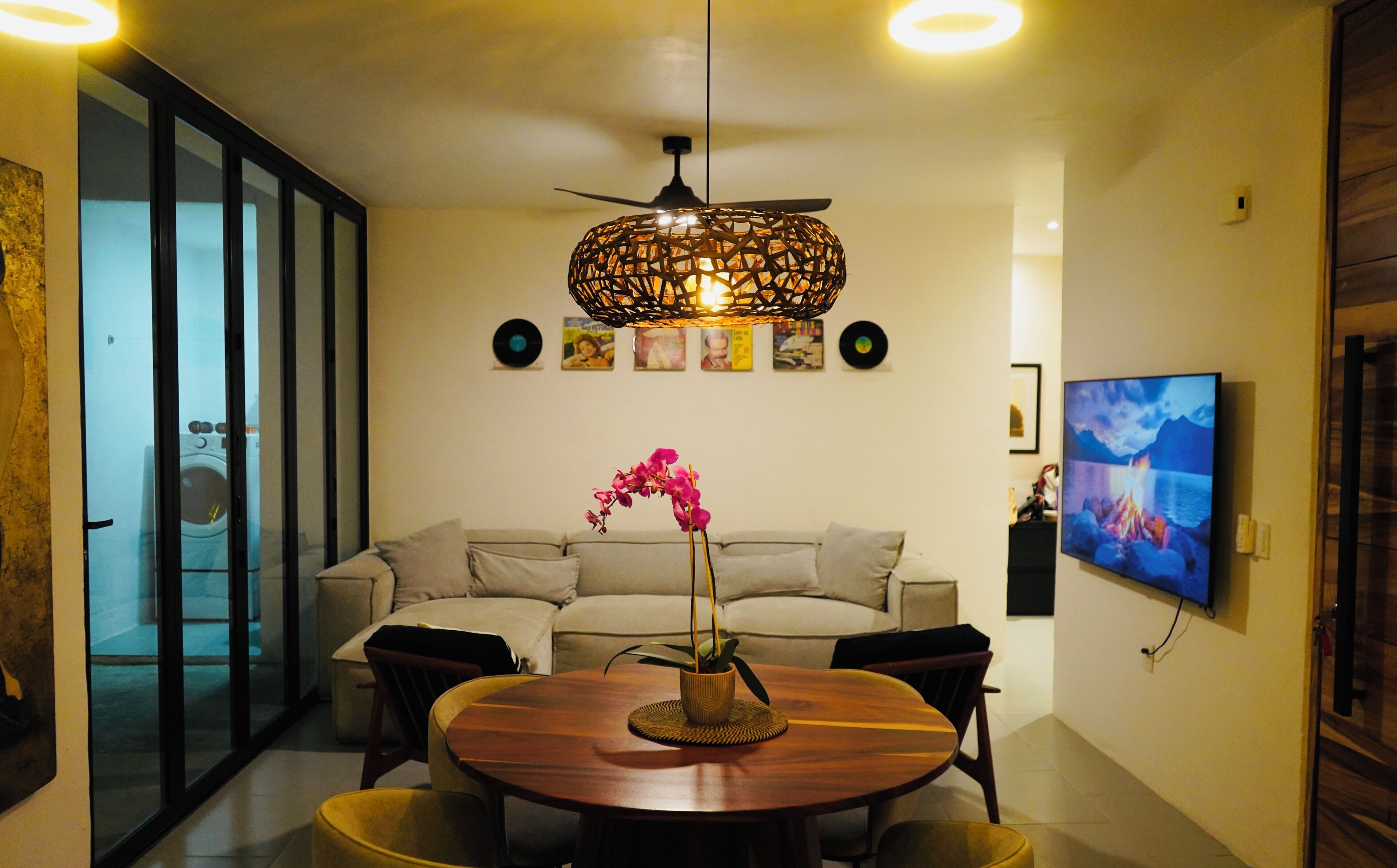 A modern living room with a round wooden dining table, a sofa, a ceiling fan, and a television mounted on the wall.