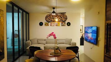 A modern living room with a round wooden dining table, a sofa, a ceiling fan, and a television mounted on the wall.