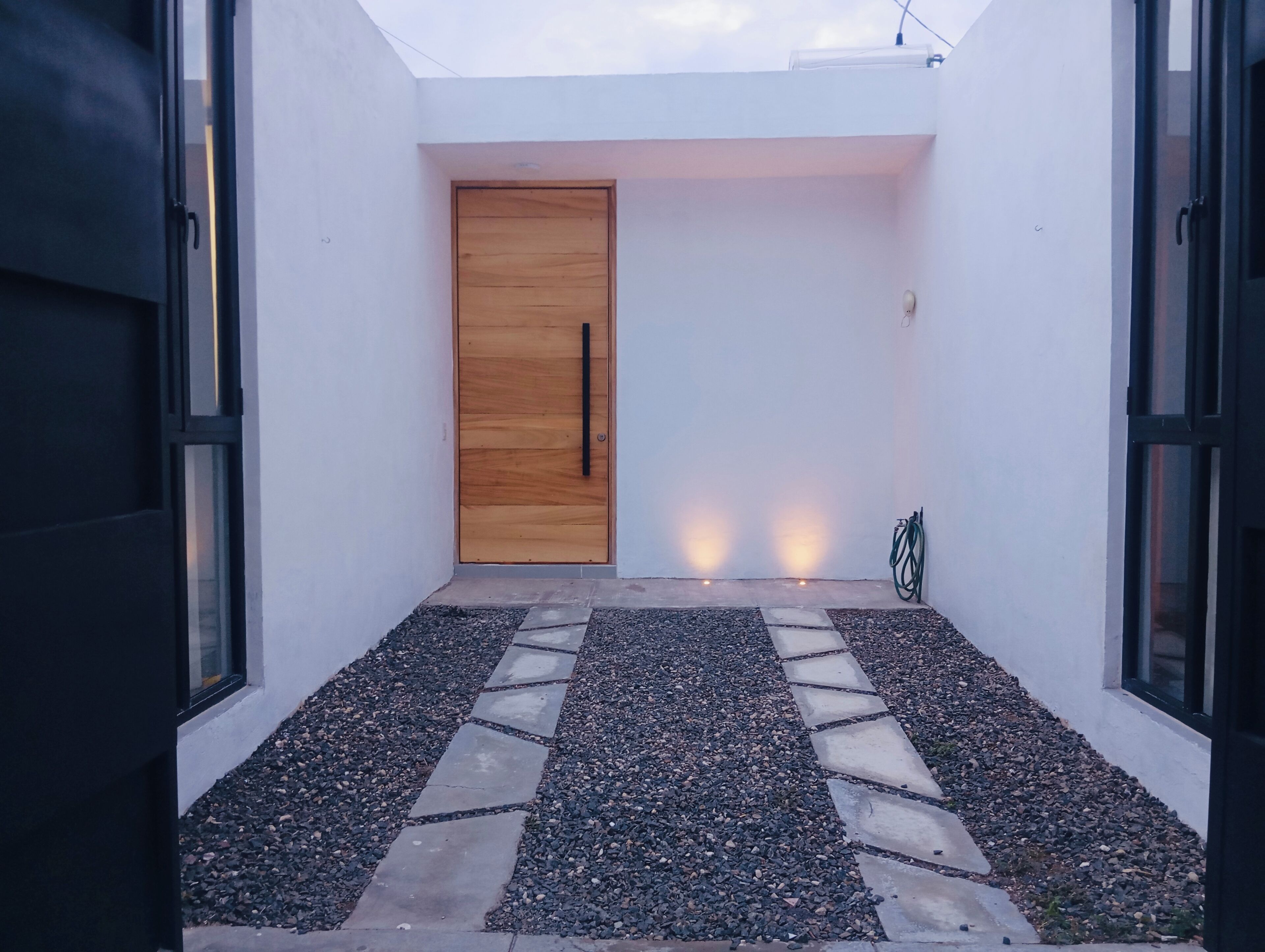 A modern entrance with a wooden door, a gravel pathway, and a hose on the right.