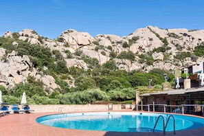 Ein Pool mit einer Steinwand und Liegestühlen, umgeben von felsigen Hügeln und viel Grün.