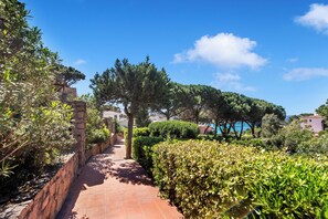 Un chemin pavé, bordé de verdure et de murs de pierre, mène à un bâtiment d’où l’on peut admirer la mer.