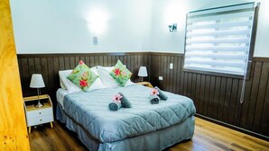 A double bed with a grey duvet, two pillows with floral patterns, and rolled towels on the bed. There is a bedside table with a lamp, a window with blinds, and a wall-mounted light fixture. - Hotel Y Cabanas Henua Iti (Hanga Roa)