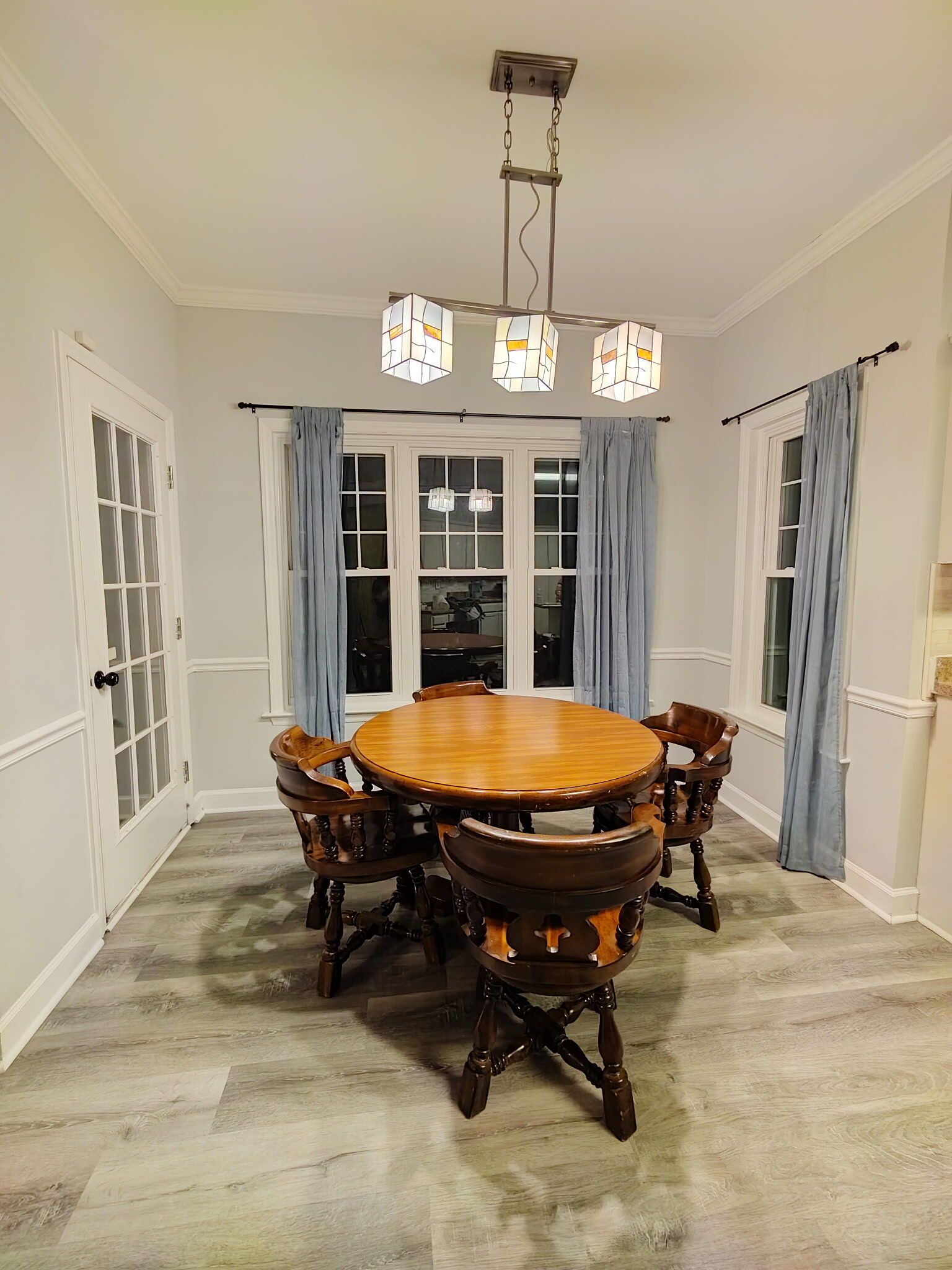 A dining room with a round wooden table and chairs, a chandelier, and a view through large windows with curtains.