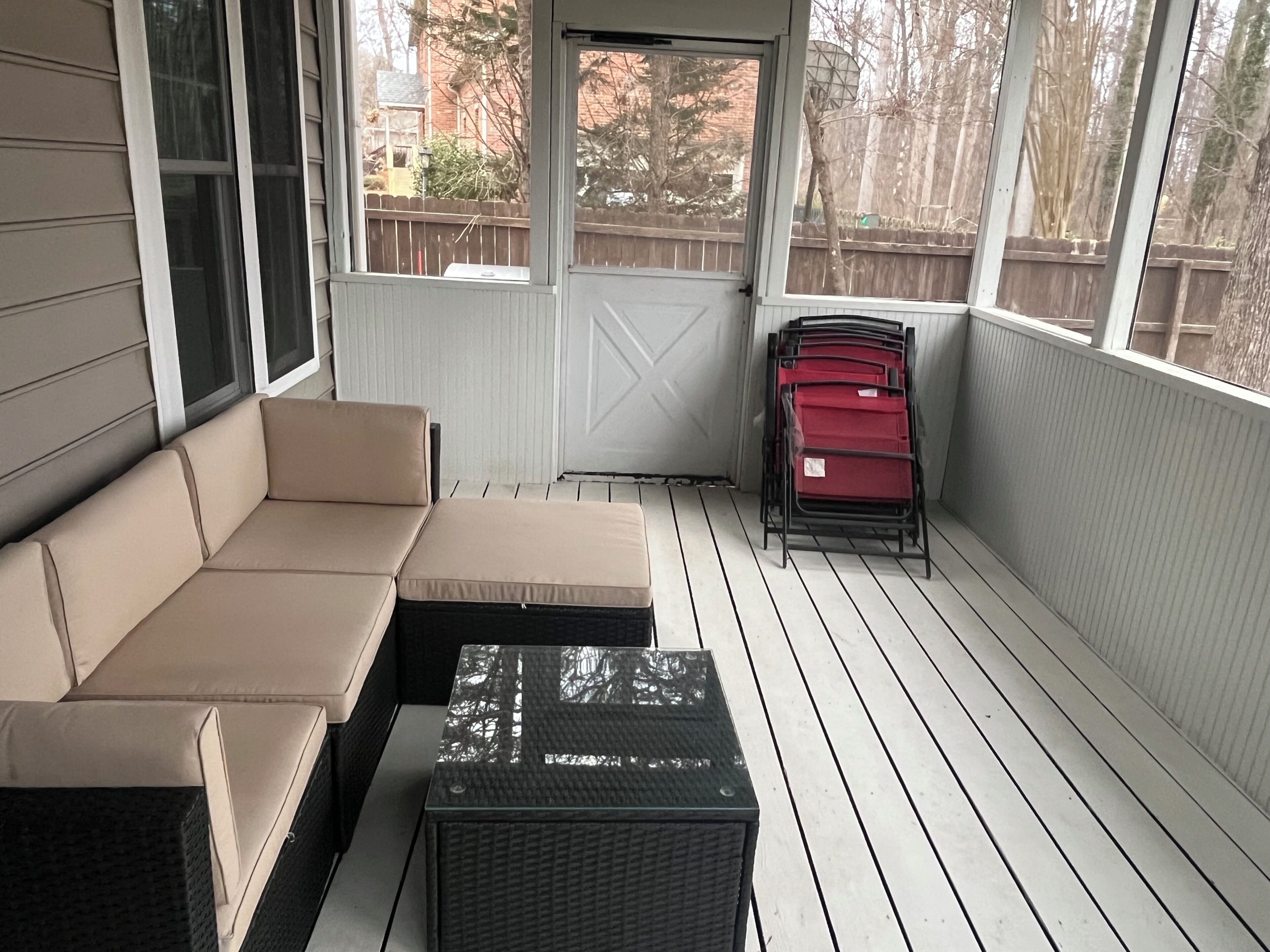 A covered porch with a beige sectional sofa, a wicker table, and a stack of red chairs.