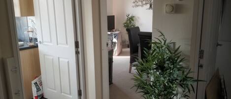 Un pasillo con una puerta blanca, una planta, un comedor con mesa y sillas, y una pared decorada.