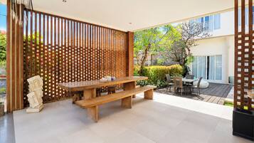 A modern outdoor dining area with a wooden table and benches, surrounded by a wooden fence and a view of a building.