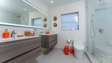 Un baño moderno con un espejo grande, ducha, inodoro y tocador con lavamanos y espacio de almacenamiento.
