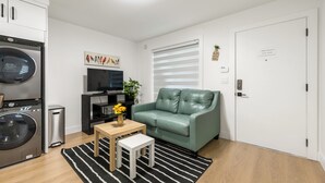 Um espaço compacto com máquina de lavar, televisão, sofá e uma pequena mesa com um vaso de flores.