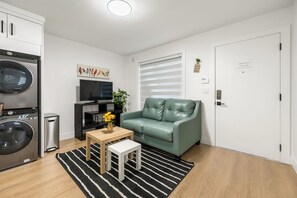 Un espacio habitable compacto con lavadora, televisor, sofá y una pequeña mesa con un jarrón de flores.
