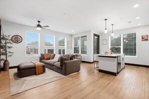 A modern living room with a sectional sofa, a small kitchen area, and large windows with blinds.