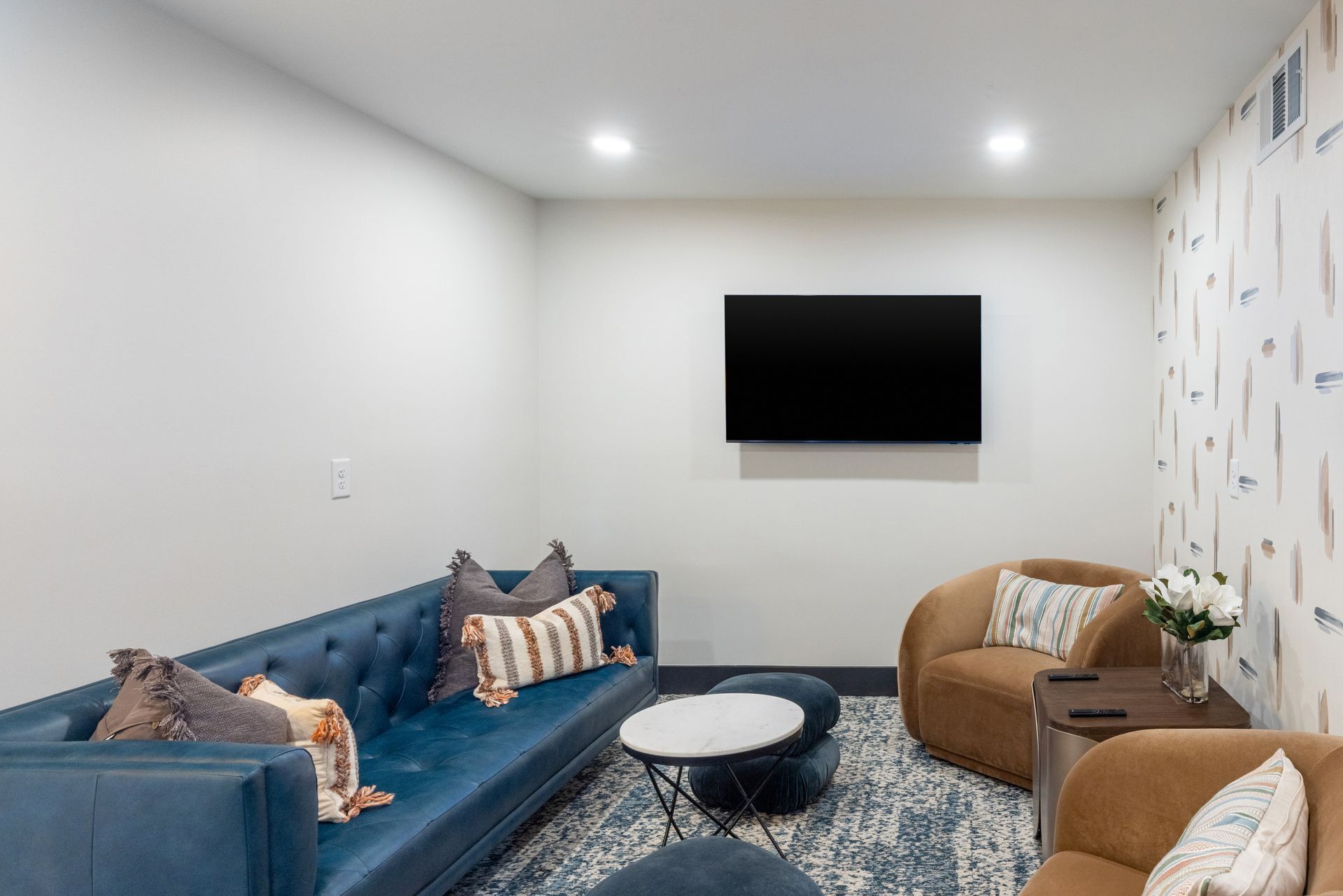 A living room with a blue sofa, a brown sofa, a round coffee table, and a flat-screen TV mounted on the wall.