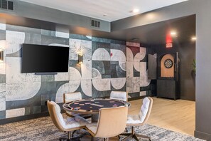 A room with a poker table, chairs, a dartboard, and a flat-screen TV mounted on the wall.
