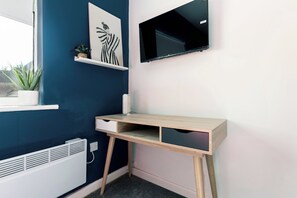 A corner room with a desk, a mounted TV, a radiator, and a potted plant.