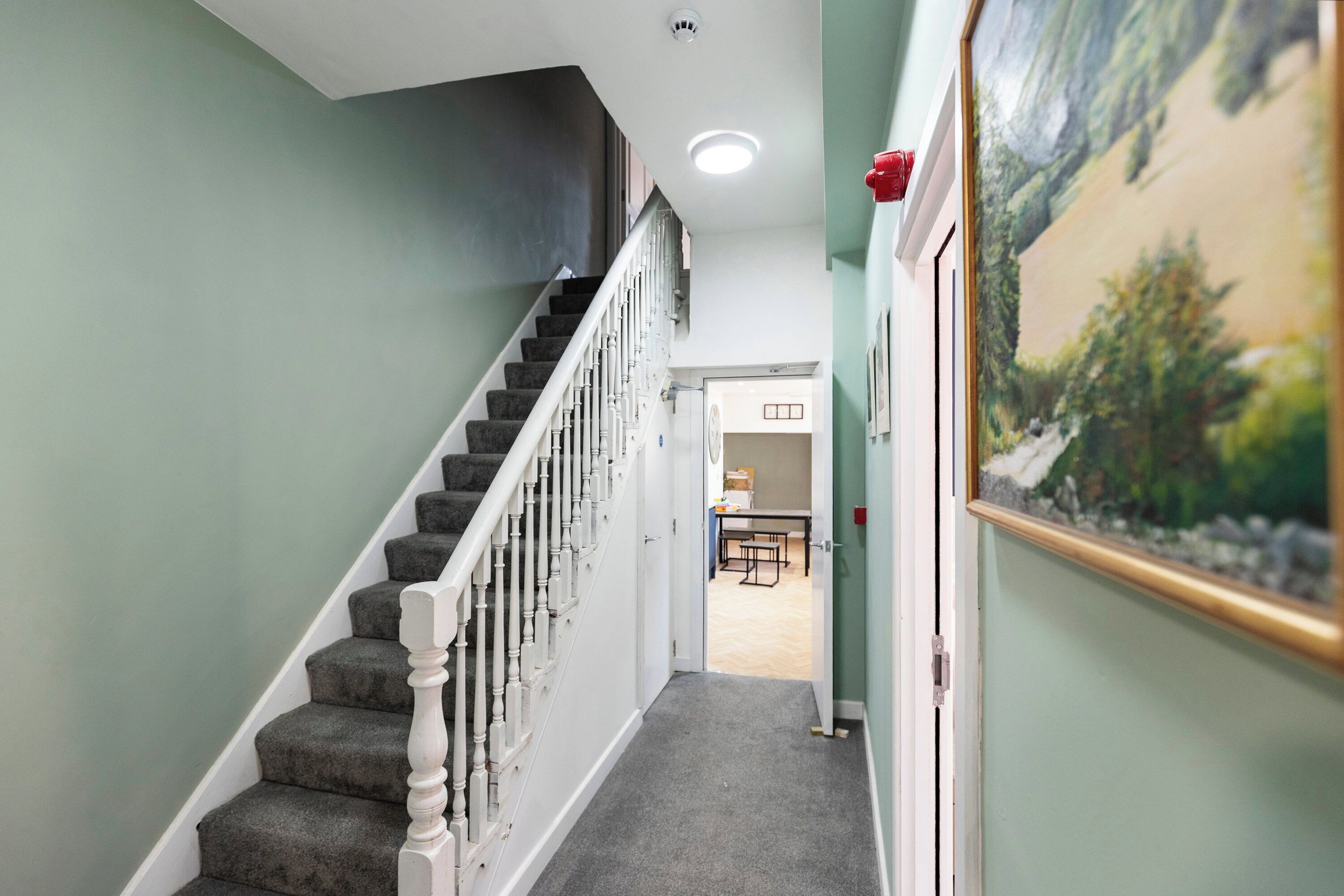 A staircase with carpeted steps and white balusters, leading to a room with a painting on the wall.