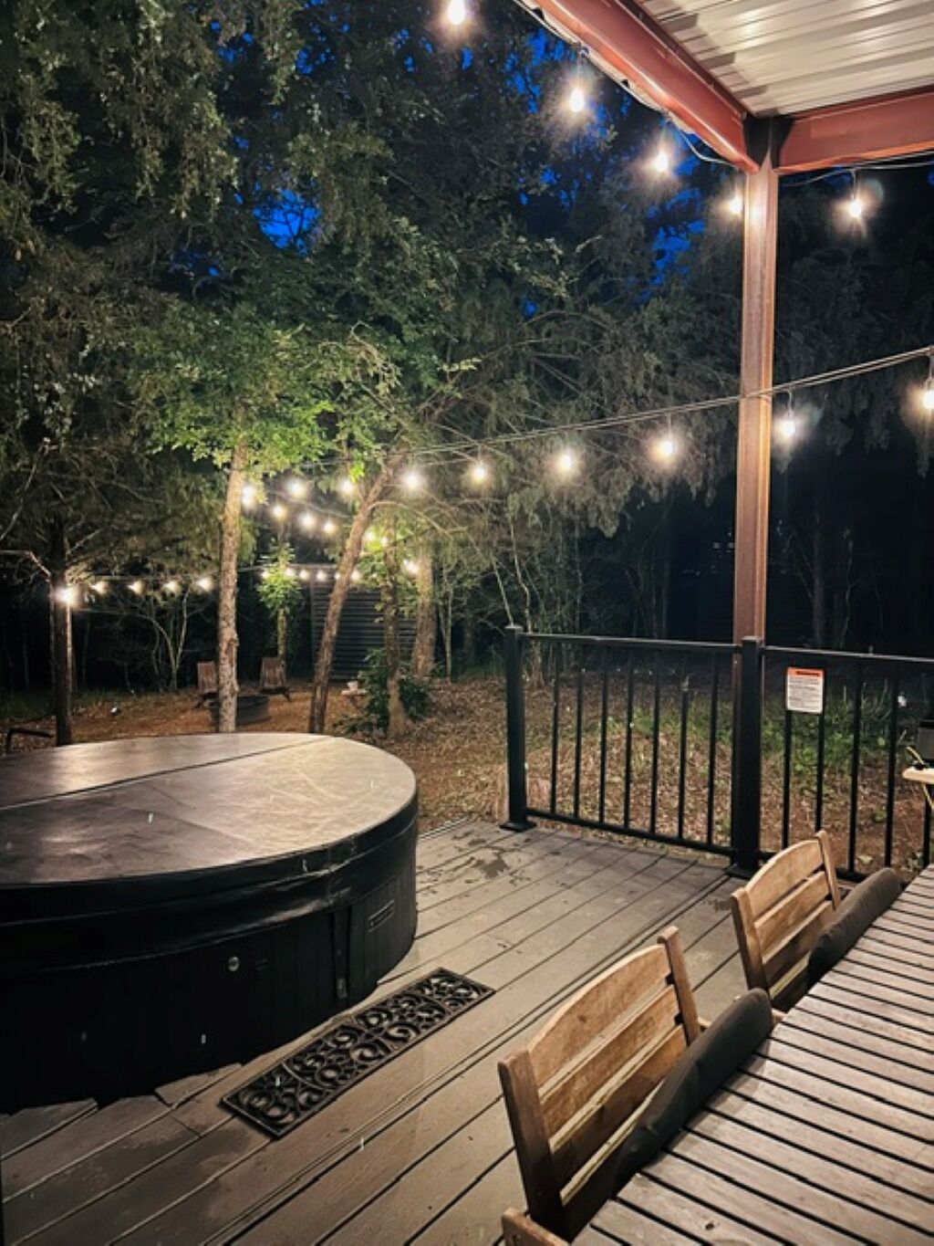 A covered outdoor deck with a hot tub, wooden benches, and string lights.