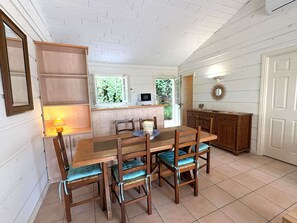 A dining area with a wooden table and chairs, a kitchen area with cabinets, and a window with greenery visible outside. - Maison climatisée dans domaine avec piscine à Gassin (GASSIN)