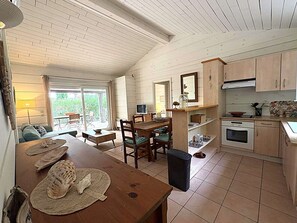A cozy, well-lit room with a dining table, chairs, a small kitchen area, and a window providing a view of greenery outside. - Maison climatisée dans domaine avec piscine à Gassin (GASSIN)