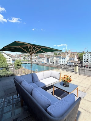 Eine Terrasse mit einer Couch, einem Tisch und einem Sonnenschirm mit Blick auf eine Stadtlandschaft und ein Gewässer.