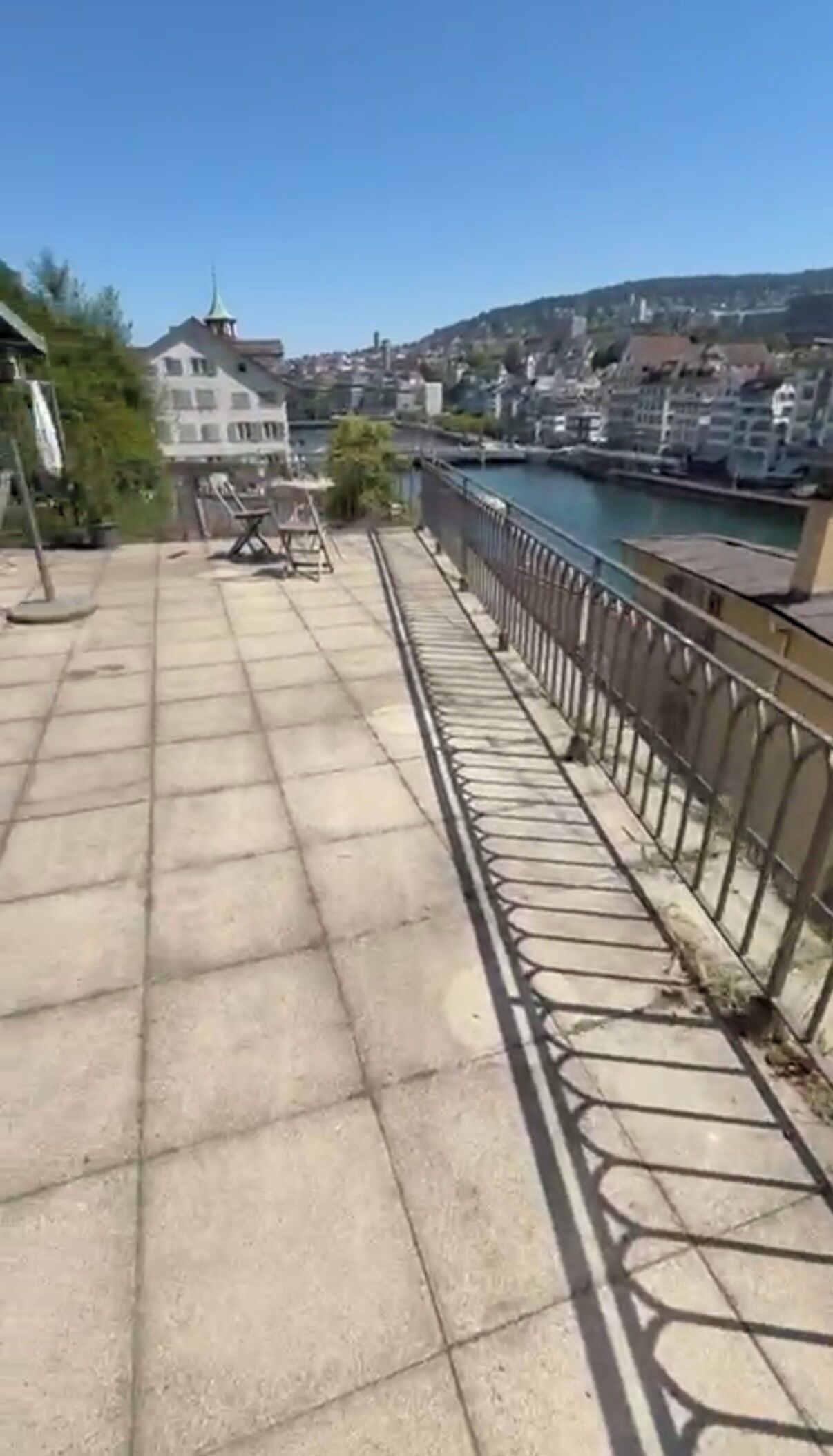Une terrasse dallée, agrémentée d’une rambarde en métal, qui offre une vue sur une rivière et des bâtiments.