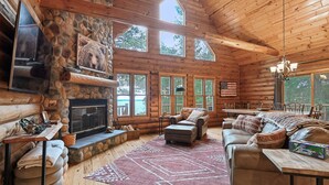 L’intérieur d’une cabane en rondins, avec une cheminée en pierre, des meubles en bois et de grandes fenêtres.