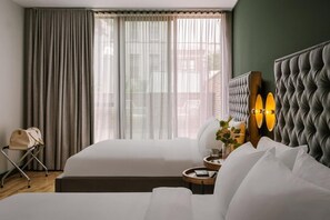 Ein Hotelzimmer mit einem großen Bett, einem Nachttisch mit Lampe, einem Sessel und Blick durch leichte Vorhänge nach draußen.