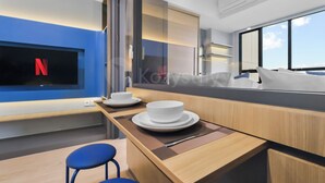 Una camera d'albergo moderna con una grande finestra, una TV a schermo piatto montata a parete, un tavolo da pranzo in legno con sottopiattino e due sedie blu.