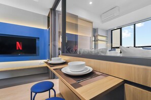 Ein modernes Hotelzimmer mit großem Fenster, einem an der Wand befestigten Flachbildfernseher, einem Holztisch mit Unterlage und zwei blauen Stühlen.