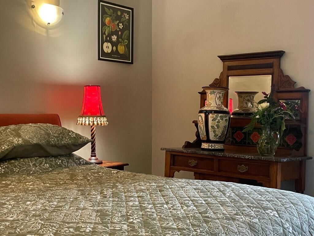 A bedroom with a bed, a nightstand with a lamp, a dresser with decorative items, and a wall-mounted mirror.