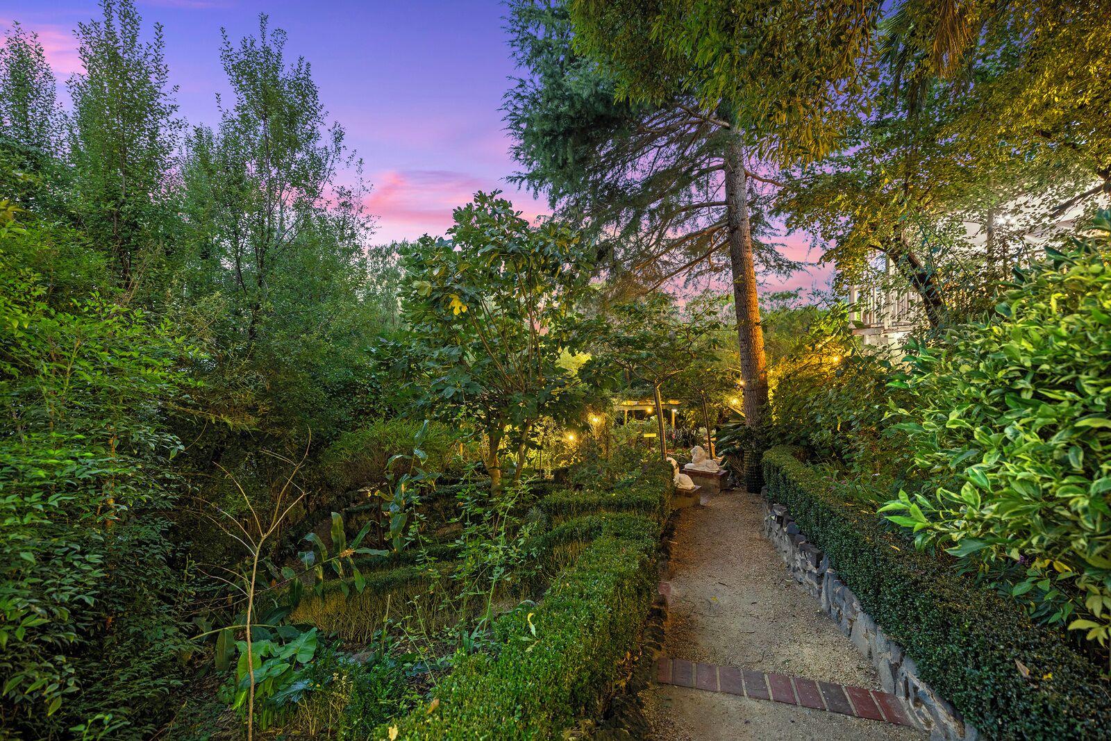 A garden pathway surrounded by dense foliage, leading to a structure with string lights.