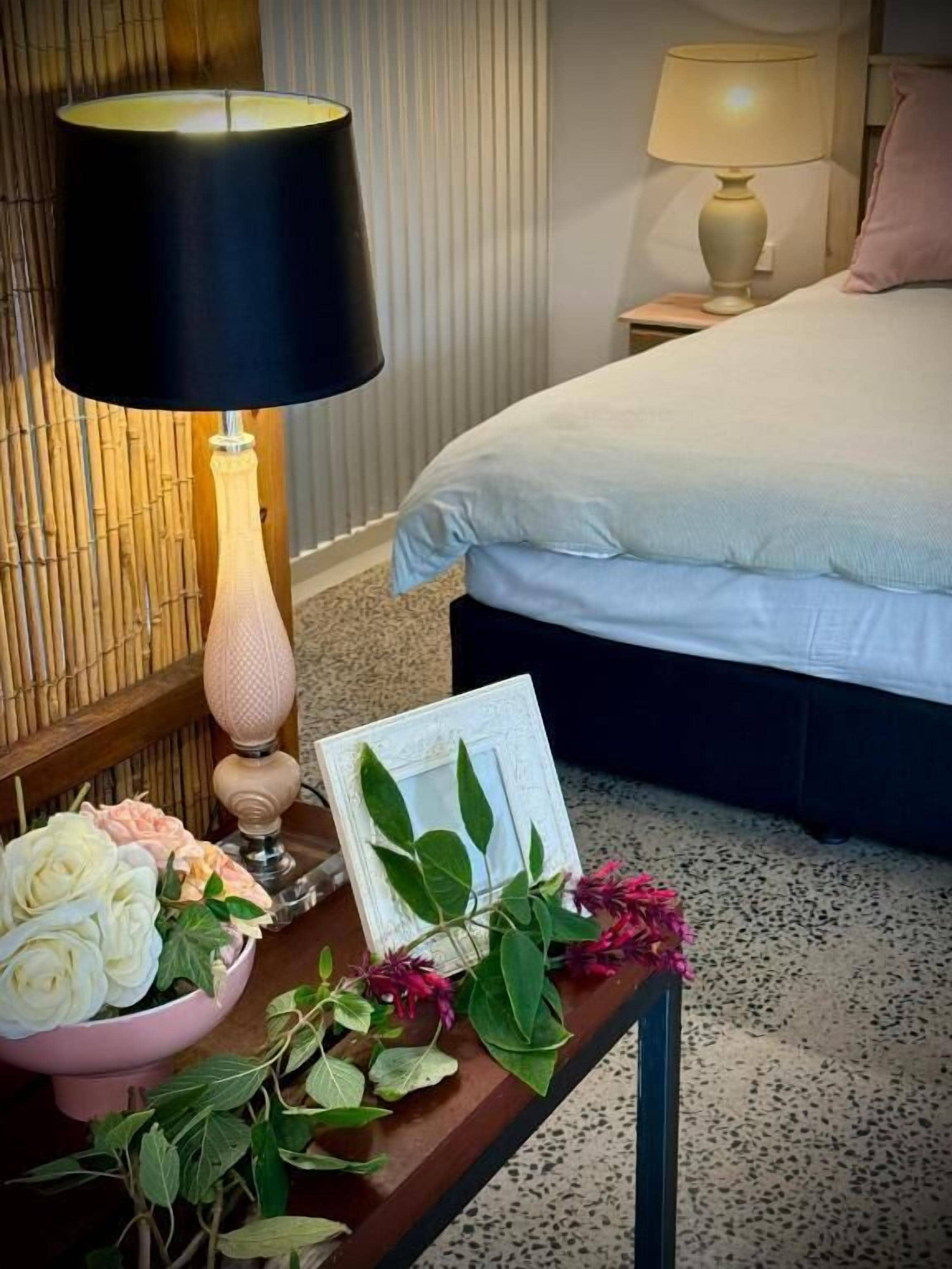 A neatly arranged bedroom with a bed, bedside lamps, a bedside table with a picture frame, and a bowl of flowers.