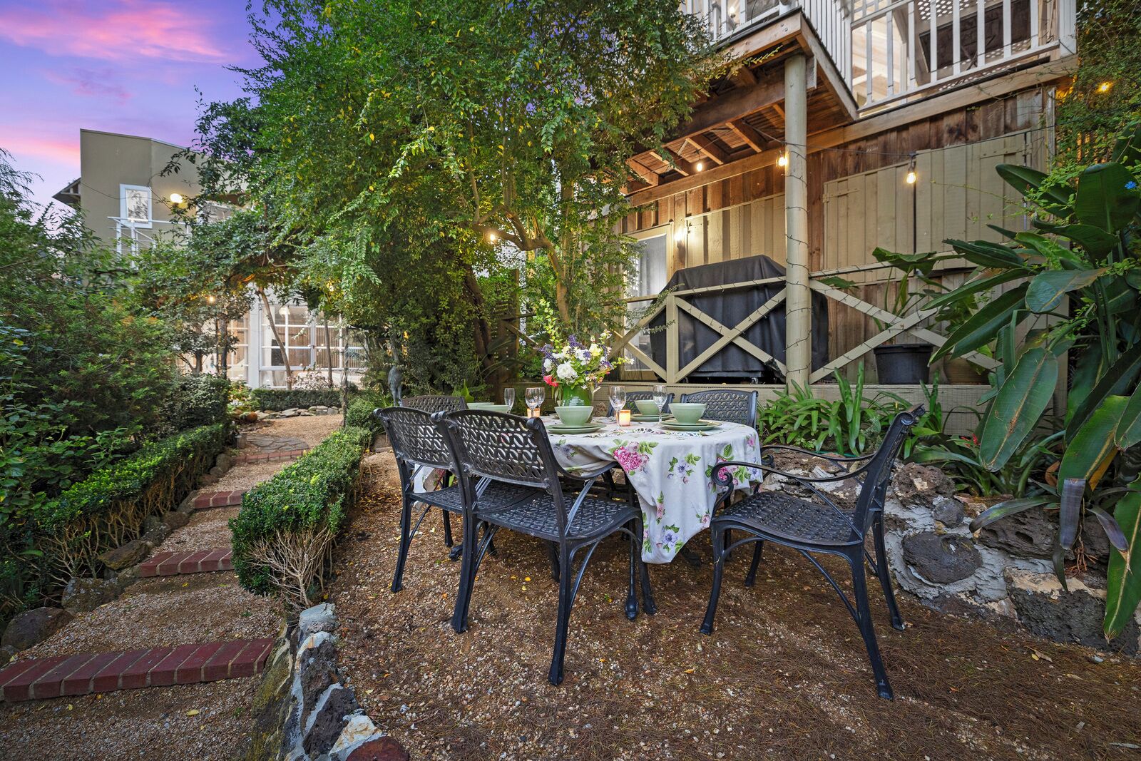 An outdoor patio with a table set for two, surrounded by greenery and a wooden fence.