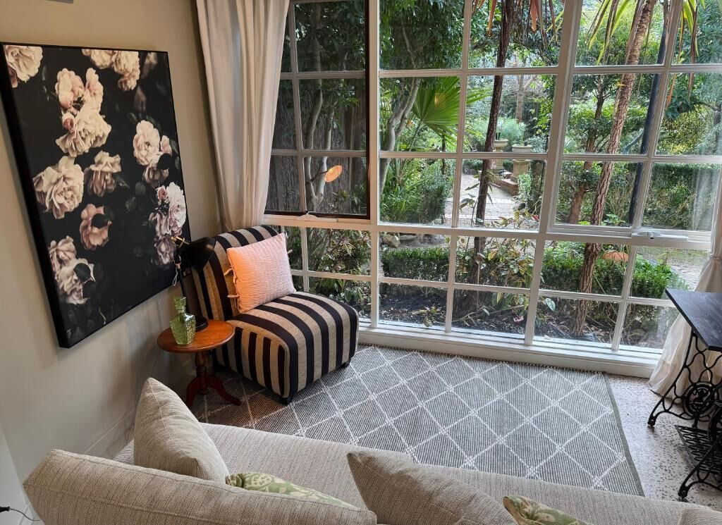 A room with a large window, a striped ottoman, a green vase, and a painting on the wall.