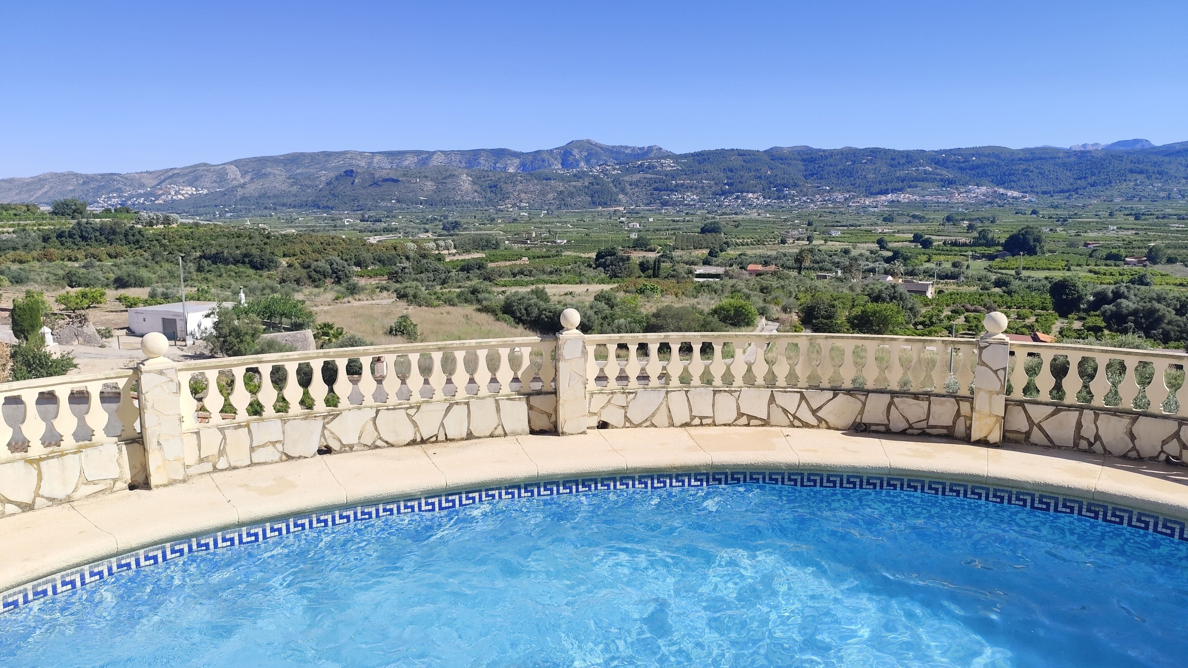 A swimming pool with a stone balustrade and a view of a rural landscape with houses and mountains.